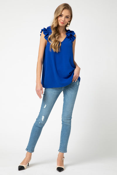 Full view of royal blue top for fall with v-neck and sleeveless ruffle detail paired with jeans and heels. Longer length for coverage.