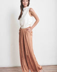 Side view of ivory satiny sleeveless top with draped neck paired with pleated satin pants and heels.