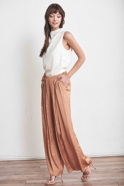 Side view of ivory satiny sleeveless top with draped neck paired with pleated satin pants and heels.